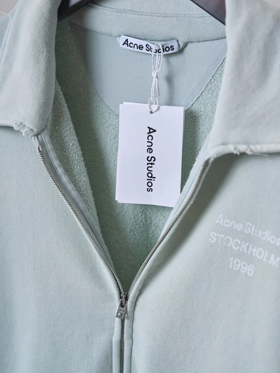 [Real auction] Acne Studios Retro distressed letter print zipper jacket [In Stock] Logo stamp at front, side-seam pockets, and ribbed cuffs and hem. Regular fit, just above the waist. It adopts a 3-strand composite fleece structure with 32 counts for the top layer and 40 counts for the bottom layer, with a weight of g/㎡. The 32-count yarn on the top layer is fine and smooth, giving the surface a smooth touch and excellent anti-pilling properties; the 40-count 3-ply yarn on the bottom layer is treated with a raising process to form a rich and soft fluff layer, providing excellent warmth and skin-friendly wrapping. With 21 2×2 ribs, the g/㎡ weight ensures that the collar, cuffs and hem have excellent tensile fatigue resistance and shape retention capabilities. During piece dyeing, the entire fabric is immersed in the dye vat after weaving, and the dye molecules fully penetrate into the fibers, ensuring full, deep colors and excellent color fastness. Each batch of dyeing produces subtle differences, making the color tone of the single product unique. On this basis, the paint is sprayed irregularly on the surface of the garment through manual spray painting to create a splash-like retro effect. The distressed processing of holes causes the color to fade and mottle in parts. The ribbed trim is also treated to form slight whitening and fluffing, which visually echoes the spray-painted effect on the body, making the new clothes appear as if they have been worn for a long time and exposed to the sun. The front logo stamp uses a slurry printing process. The printing slurry contains a special microcapsule foaming agent. After being baked at high temperature, the slurry layer expands to form three-dimensional bulges, and the surface presents a granular texture similar to inkjet paint. This process simulates the effect of random drops of pigment after being atomized during spray gun spraying. The edges of the pattern present a natural feathering transition, rather than the rigid boundaries of glue printing. The printing surface has a fine graininess and soft elasticity to the touch, forming a closed loop of the process with the distressed effect of the spray painting on the body.